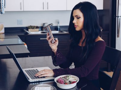 Woman sitting at table with her laptop and a salad while holding her phone