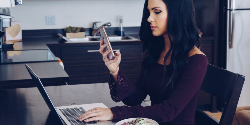 Woman sitting at table with her laptop and a salad while holding her phone